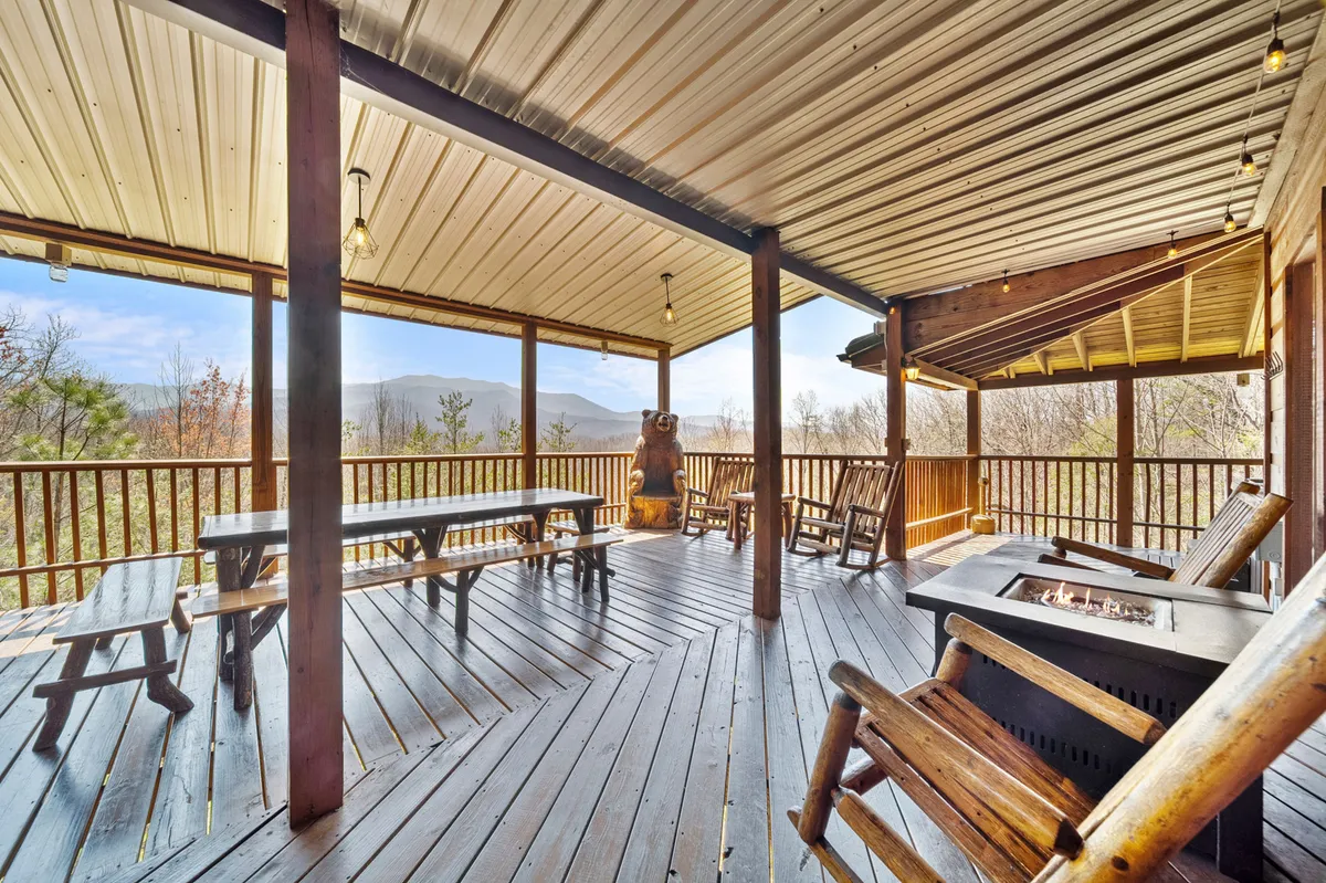 A spacious deck featuring multiple rocking chairs, a fire pit, picnic tables, and expansive mountain views. The scene captures sunset lighting and a communal, cozy atmosphere, ideal for gatherings, outdoor dining, and storytelling by the fire.