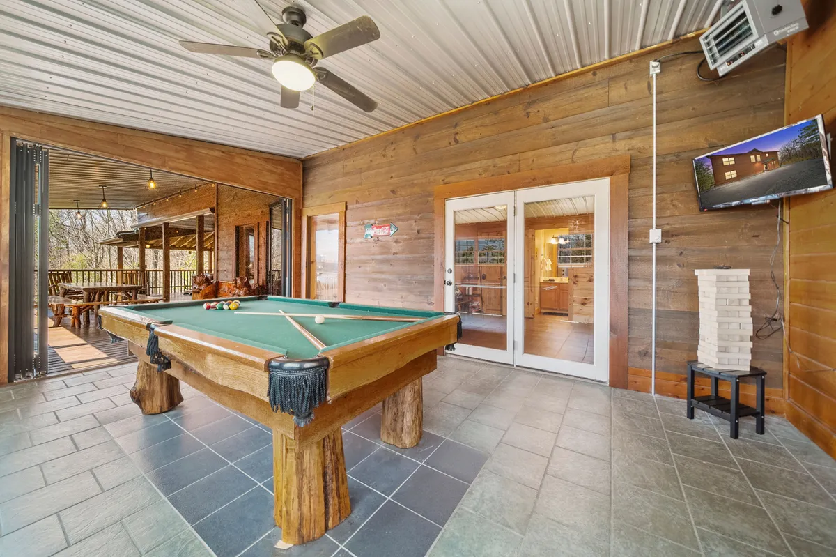 A welcoming pool table room with rich wooden walls, a pool table ready for play, a wall-mounted television, and sliding doors that let in natural light. Bar-style seating encourages socializing, and the room feels geared for both friendly competition and relaxation.