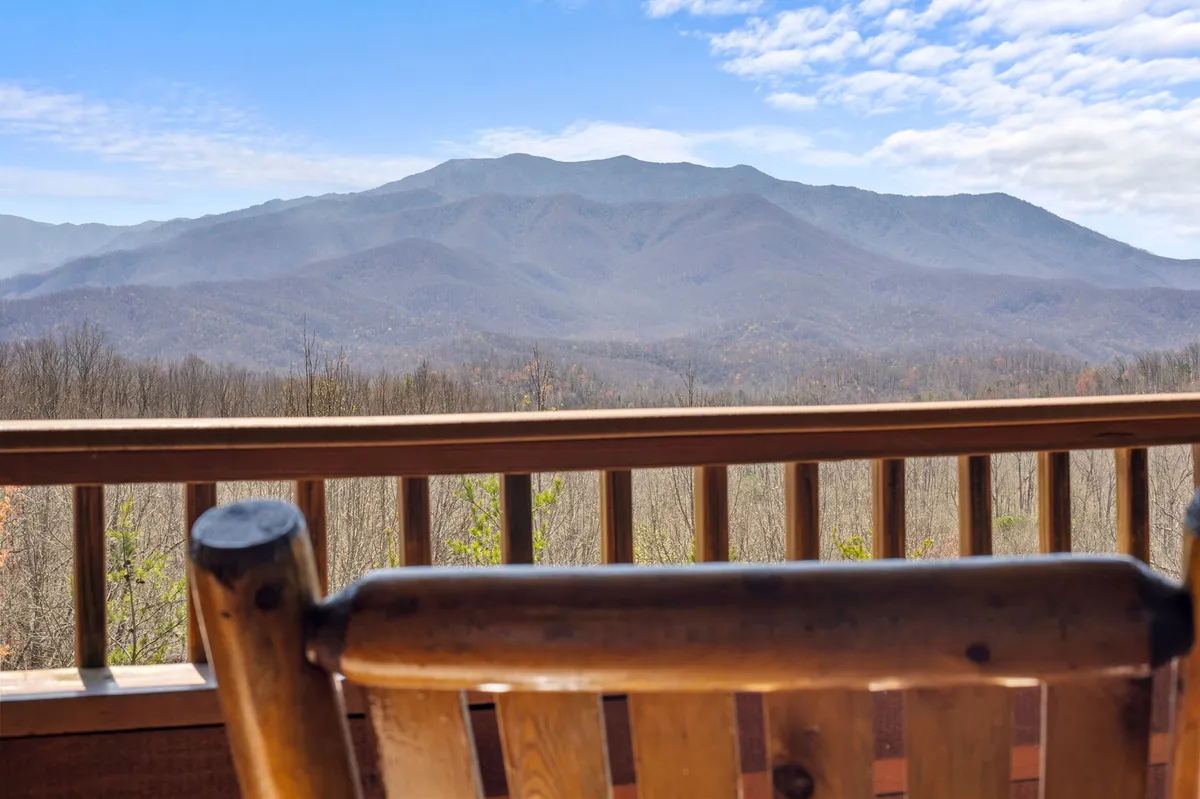 This image captures a breathtaking mountain range beyond a wooden deck, with a classic rocking chair positioned to take in the serene views. The wooden railing frames the scenic panorama, evoking a sense of peace and immersion in nature.
