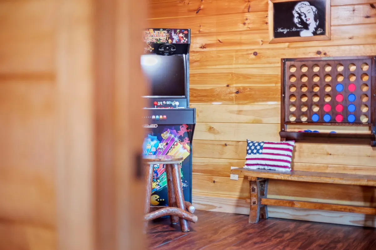 The image captures a vibrant game room featuring a classic arcade machine, a Connect Four board game, a wooden bench for seating, and playful decor including a Marilyn Monroe portrait and an American flag pillow. The setup invites guests to play arcade and board games or enjoy relaxed conversations.