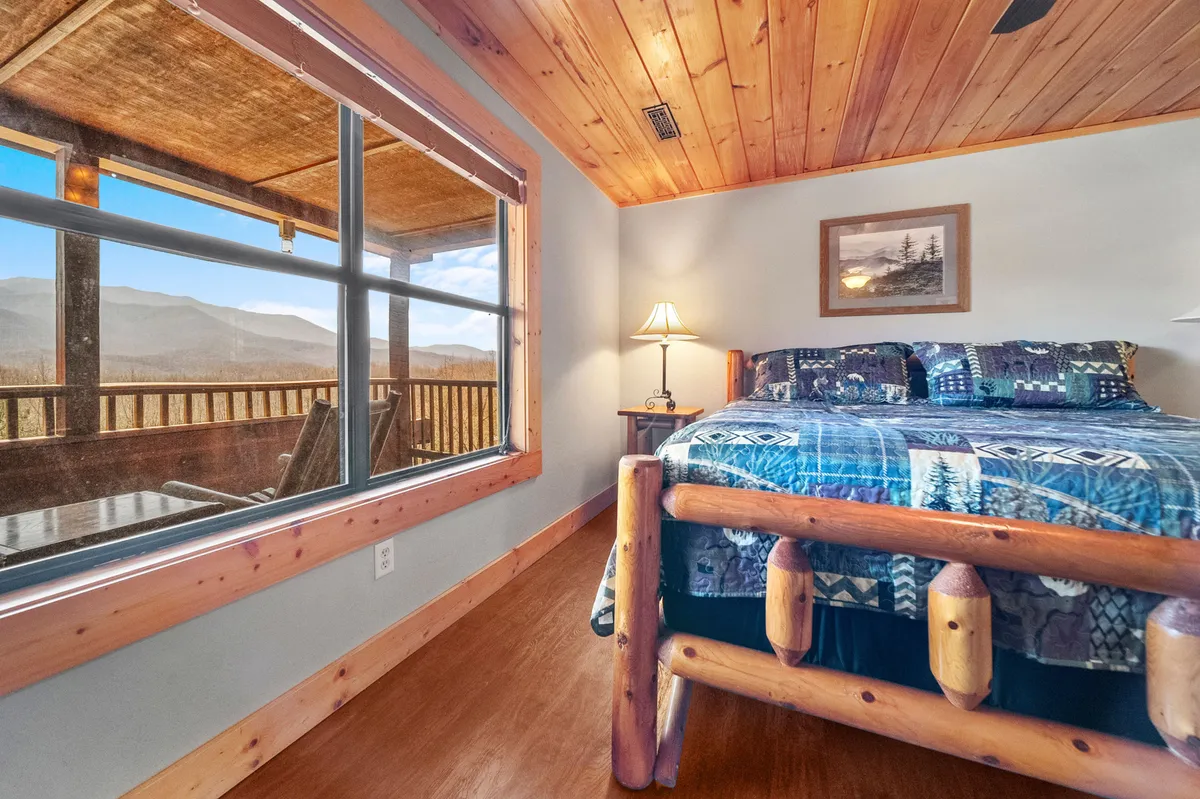A tranquil bedroom featuring a king bed with patterned bedding, a large window offering a stunning mountain view, and a cozy table lamp providing a warm ambiance. The room exudes calm and comfort, accentuated by natural light and soft furnishings.