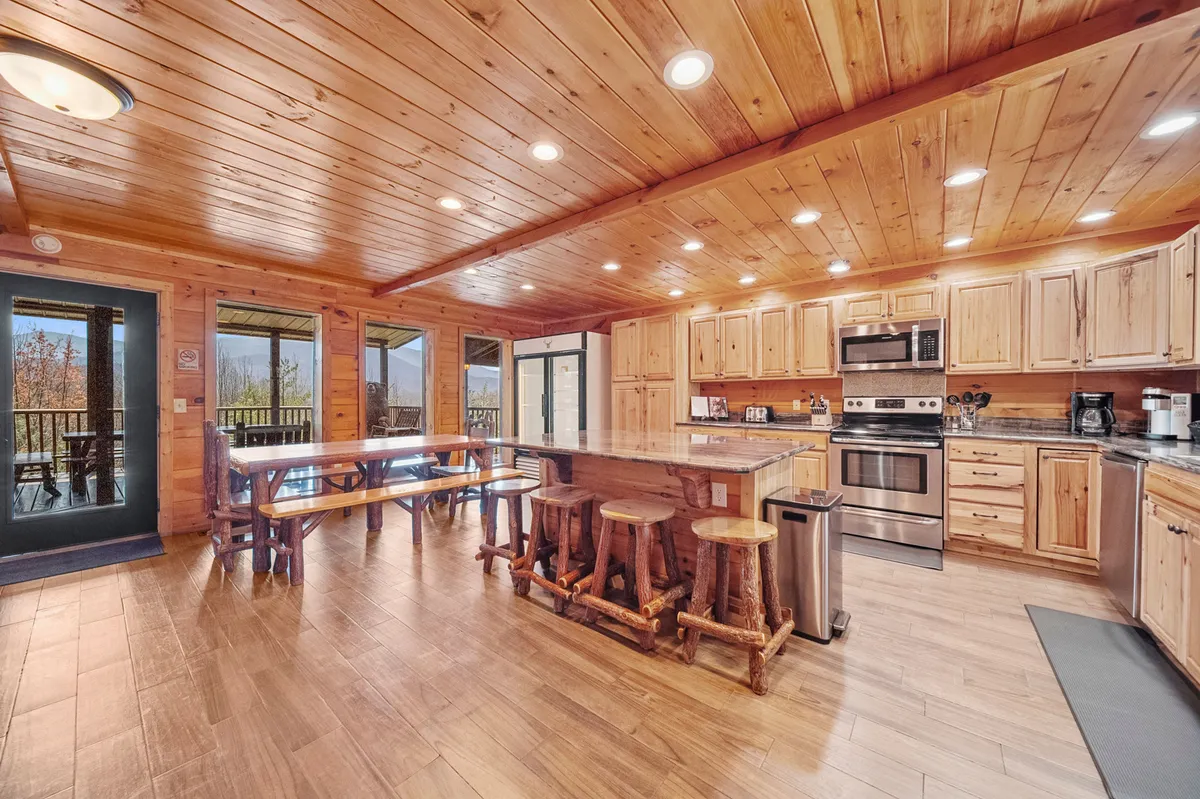 The kitchen and dining space features a large wooden kitchen island, stainless steel appliances, a spacious dining table, and panoramic windows framing scenic mountain views. The setup is ideal for group cooking and communal meals.
