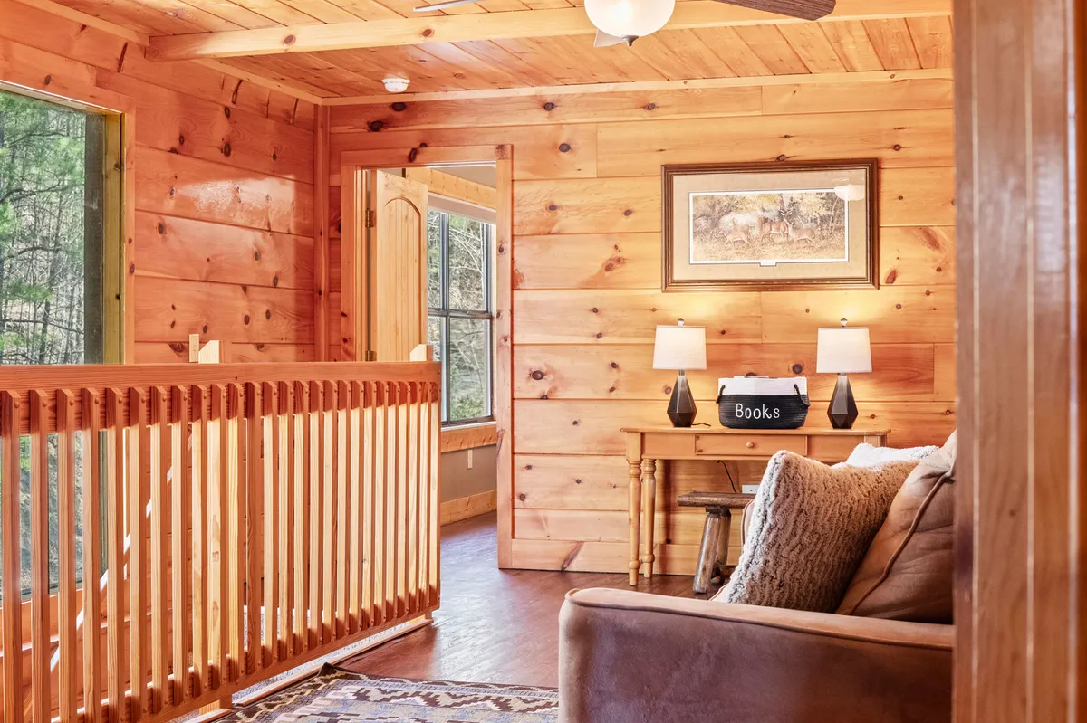 A cozy loft area furnished with a comfortable couch, wooden railing, bookshelf filled with books, and framed artwork. The space is bathed in natural light, ideal for quiet reading, reflection, or enjoying serene moments away from the main living spaces.