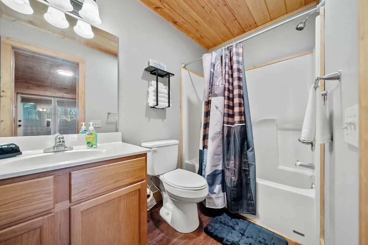 A warmly lit bathroom with a wooden ceiling, a functional vanity and sink, a neatly hung towel rack, and a decorative shower curtain. The space is designed for relaxation and personal care, with inviting finishes and practical amenities.
