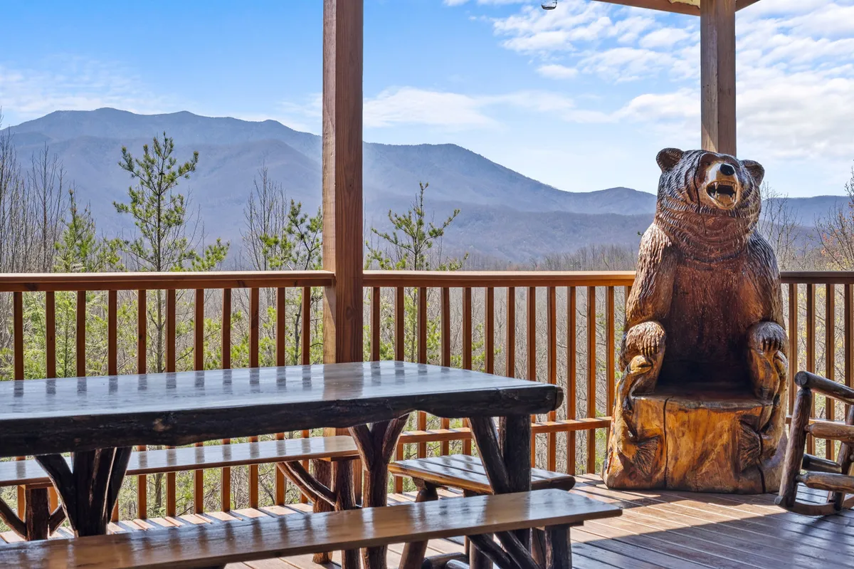 An outdoor picnic table sits beside a charming carved bear statue, all overlooking a sweeping mountain view. The setting is perfect for outdoor dining, casual gatherings, or simply savoring the scenery.